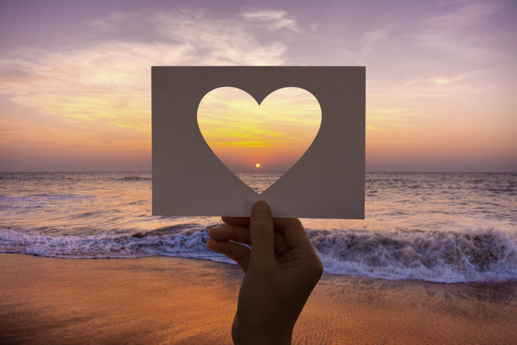 Photograph of the beach at sunset with a hand holding up a paper heart cutout